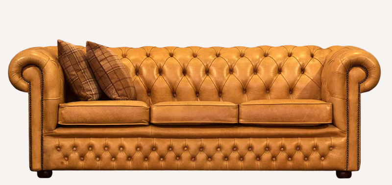 A classic Chesterfield-style leather sofa in a warm tan/caramel color, featuring deep button tufting, rolled arms, and two brown plaid throw pillows, shown against a plain white background.