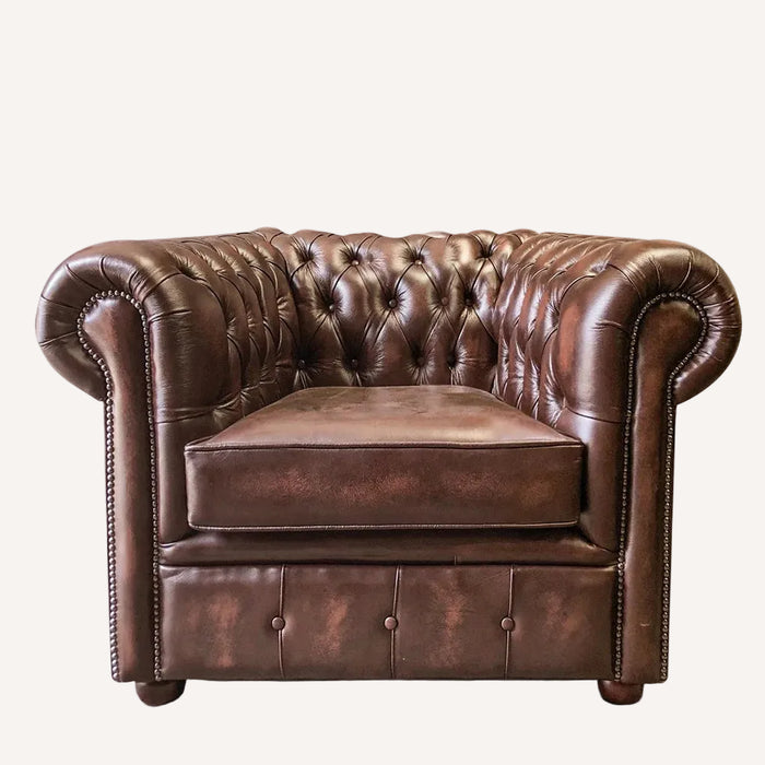 A classic Chesterfield armchair in distressed brown leather, featuring deep button tufting on the backrest, rolled arms, nailhead trim, and a matching seat cushion, shown against a plain white background.
